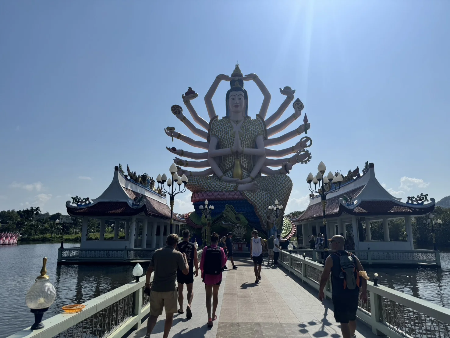 Large multi-armed Guanyin statue at Wat Plai Laem temple in Koh Samui, Thailand, with visitors walking along the entrance bridge.