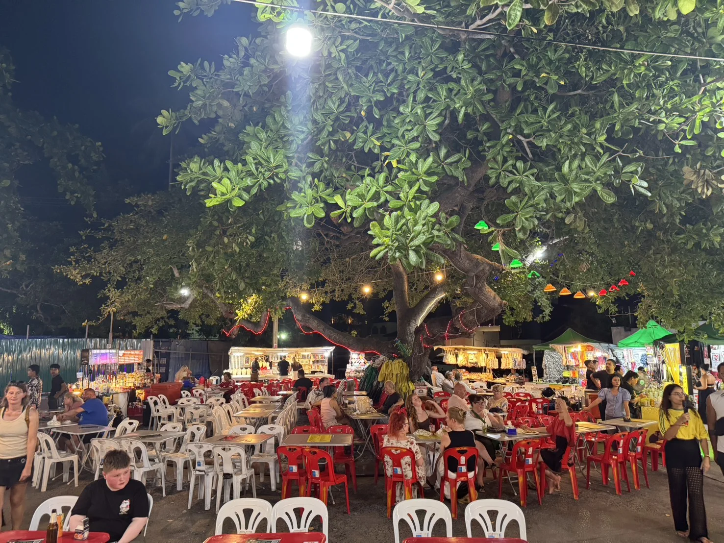 Outdoor dining area at Fisherman's Village in Bophut, Koh Samui, with colorful tables, evening lights, and restaurants beneath a large tree.