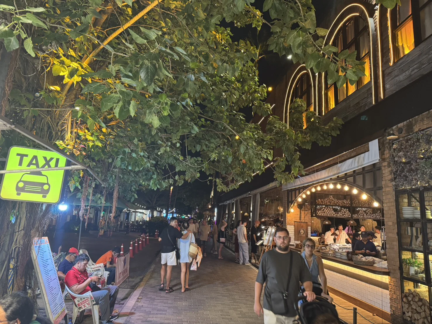Evening street scene at Fisherman's Village in Bophut, Koh Samui, with illuminated restaurants, shops, and visitors walking beneath the trees.
